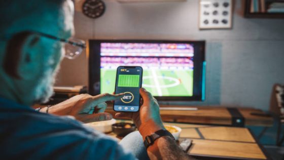 Mature men sitting on sofa at home and watching soccer game. He is using smart phone for sports betting .
